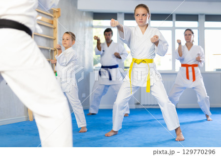 Teen girl with her family in kimonos and colored belts practicing karate with punches during group martial arts class in gym, accompanied by trainer Teen girl with her family in kimonos and colored belts practicing karate with punches during group martial arts class in gym, accompanied by trainer 129770296