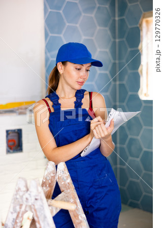 Woman in blue overalls checking completed construction work on drawing 129770326