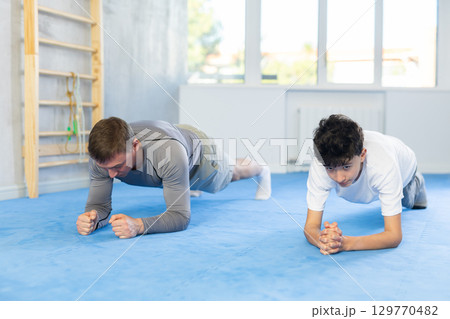 Underage boy and middle-aged trainer practicing plank pose in sports hall 129770482