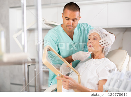 Male plastic surgeon giving professional advice after facial contouring to senior woman client looking at mirror Male plastic surgeon giving professional advice after facial contouring to senior woman client looking at mirror 129770613