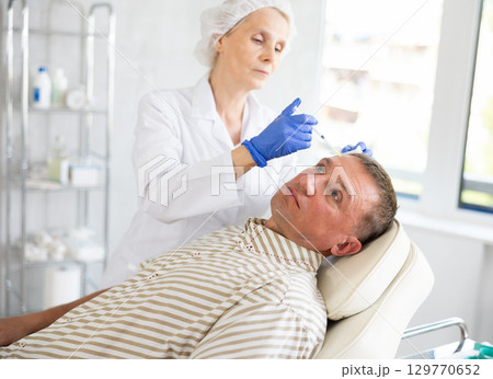 Mature woman doctor gives injection and performs procedure of lip augmentation of male patient Mature woman doctor gives injection and performs procedure of lip augmentation of male patient 129770652