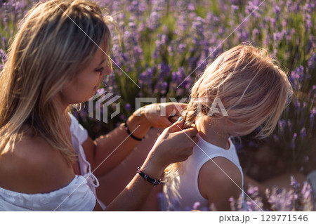 Mother Daughter Lavender: Woman braids girl's hair amidst purple flowers during a sunny day bonding. Mother Daughter Lavender: Woman braids girl's hair amidst purple flowers during a sunny day bonding. 129771546