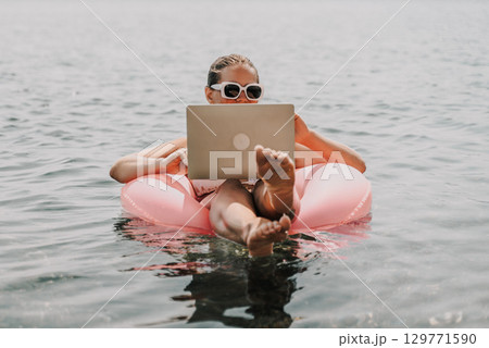 A woman is floating in a pink inflatable raft with a laptop in her hand. She is smiling and she is enjoying her time in the water. 129771590