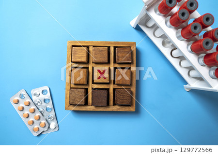 Closeup view of medicine tablet pills and a test tube holder with wooden tic tac toe game on a blue background. Concept of healthcare and medicine Closeup view of medicine tablet pills and a test tube holder with wooden tic tac toe game on a blue background. Concept of healthcare and medicine 129772566