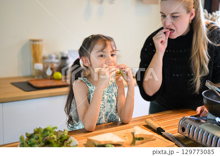 Happy mother and daughter eating sandwiches together in the kitchen, enjoying a homemade breakfast Happy mother and daughter eating sandwiches together in the kitchen, enjoying a homemade breakfast 129773085