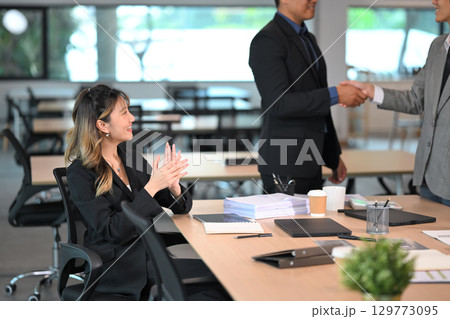 Two businessmen shaking hands in a modern office while a female colleague claps in support Two businessmen shaking hands in a modern office while a female colleague claps in support 129773095