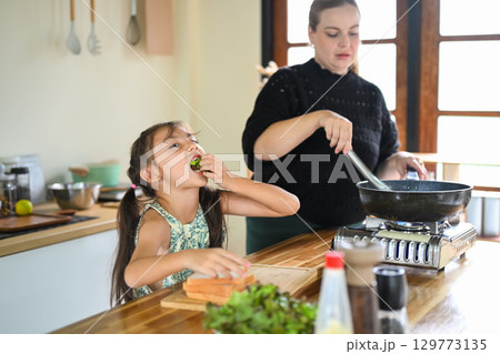 Little girl eats fresh vegetables while her mother cooks breakfast in the kitchen Little girl eats fresh vegetables while her mother cooks breakfast in the kitchen 129773135