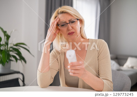 Worried blonde woman in beige clothes holding thermometer, experiencing high fever and poor health in modern apartment living room Worried blonde woman in beige clothes holding thermometer, experiencing high fever and poor health in modern apartment living room 129773245