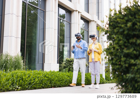 A man and a woman wearing VR headsets are walking outside a modern building. They are both exploring virtual reality. A man and a woman wearing VR headsets are walking outside a modern building. They are both exploring virtual reality. 129773689