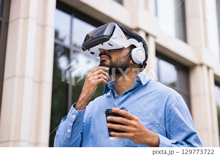 A man wearing a VR headset with coffee, looking at the building. Thinking about the future of technology and virtual reality. 129773722