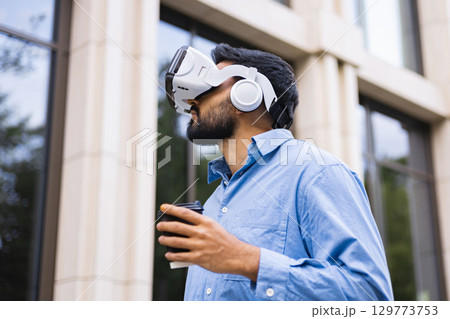 A man wearing a VR headset outside, holding a coffee cup near a building. 129773753