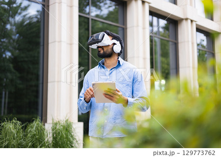 A man wearing a VR headset and headphones, holding a tablet in front of a building. 129773762