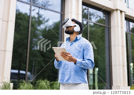 A man wearing a VR headset and holding a tablet stands outside a building, experiencing virtual reality. A man wearing a VR headset and holding a tablet stands outside a building, experiencing virtual reality. 129773813