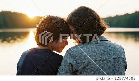 Loving mother and son touching foreheads by a serene lake at golden hour sunset. 129774505