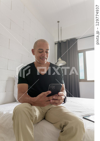 Bald Hispanic man sitting on bed at home using phone Bald Hispanic man sitting on bed at home using phone 129774937