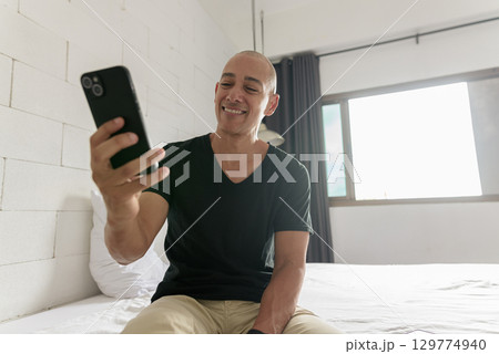 Bald Hispanic man sitting on bed at home using phone 129774940