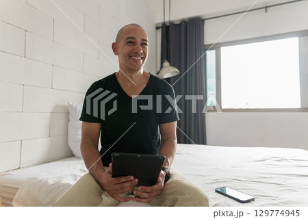 Hispanic bald man sitting on bed while using digital tablet computer smiling and looking away 129774945