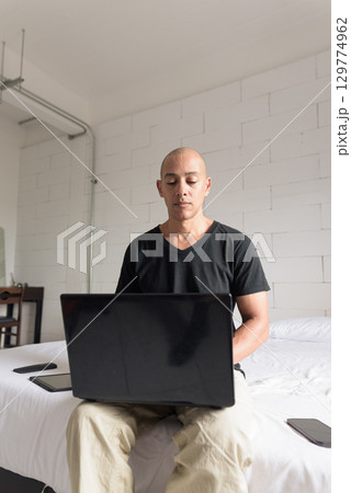Remote work from home concept bald Hispanic man sitting on bed using laptop computer 129774962