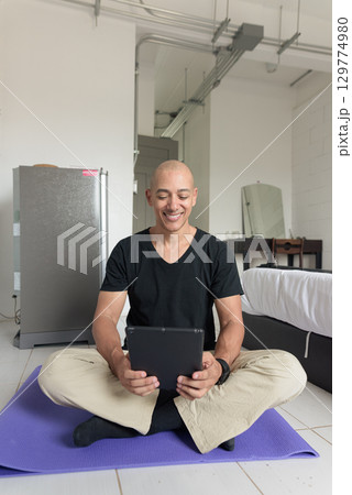 Man sitting on yoga mat doing meditation and wellness exercise at home and using tablet computer 129774980