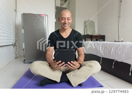 Man sitting on yoga mat doing meditation and wellness exercise at home and using tablet computer 129774983