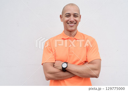 Bald Italian man in orange polo shirt studio portrait against white wall with arms crossed smiling Bald Italian man in orange polo shirt studio portrait against white wall with arms crossed smiling 129775000