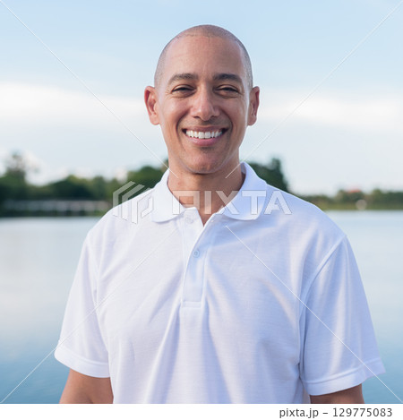 Confident bald man in white polo shirt posing by lake in serene outdoor portrait 129775083