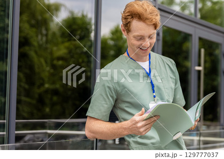 Red-haired healthcare worker in lab coat standing near glass building holding documents 129777037