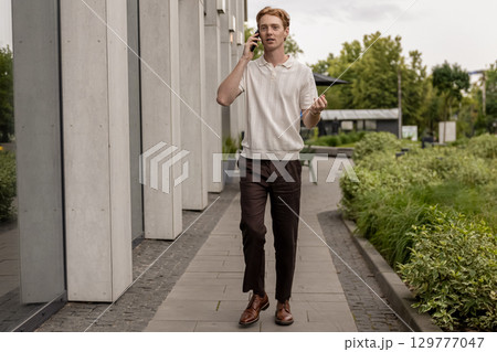 Ginger guy walking past office building and talking on mobile phone 129777047