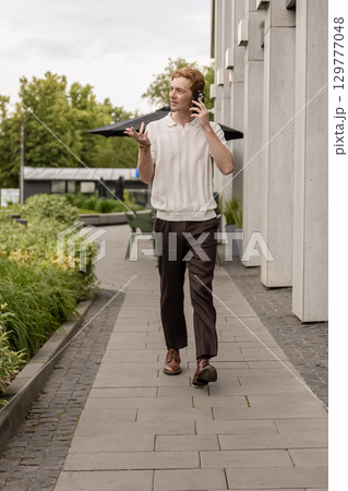 Ginger man walking along street by office building having conversation on smartphone Ginger man walking along street by office building having conversation on smartphone 129777048