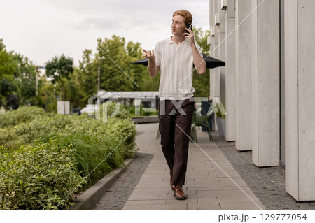 Ginger man walking along street by building and talking actively on smartphone 129777054