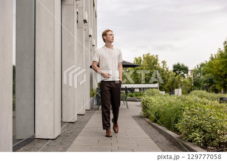 Young male with ginger hair walking outdoors holding closed laptop near modern building 129777058