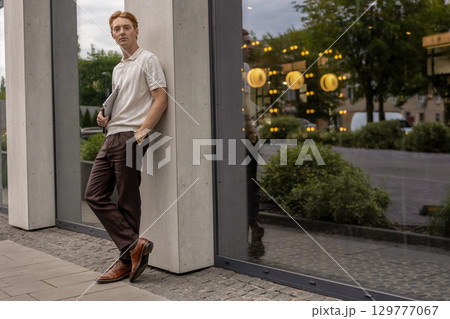 Ginger guy standing still near neutral wall holding laptop and focusing on something in distance Ginger guy standing still near neutral wall holding laptop and focusing on something in distance 129777067