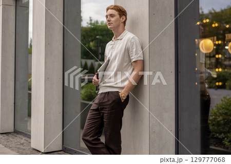 Ginger-haired young adult leaning lightly on wall holding laptop in one hand Ginger-haired young adult leaning lightly on wall holding laptop in one hand 129777068