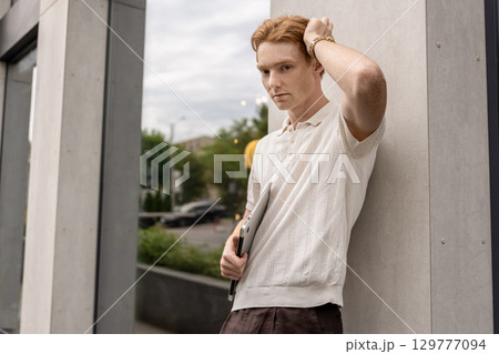 Red-haired man posing with closed laptop while standing by building wall Red-haired man posing with closed laptop while standing by building wall 129777094