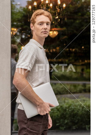Man walking past office building and passing green flower beds carrying laptop Man walking past office building and passing green flower beds carrying laptop 129777165