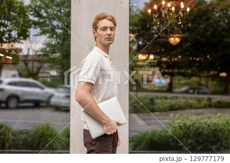 Young male with ginger hair moving along street holding laptop near office building Young male with ginger hair moving along street holding laptop near office building 129777179