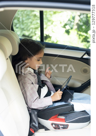 Dark-haired girl of school age sitting in a care with her seatbelt fixed 129777318