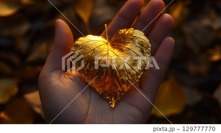 A golden autumn leaf in the shape of a heart, held in the palm of a hand, with autumn leaves scattered beneath . 129777770