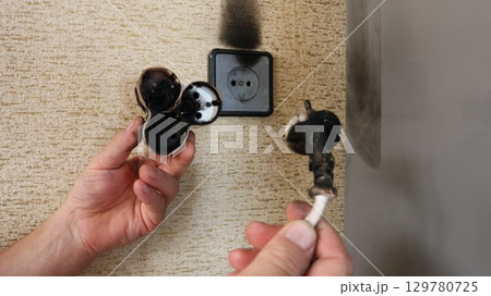 hands holding a burnt electrical plug and a scorched tee, victims of a flash and fire in a socket on the wall of a house or apartment, a dangerous situation with home electrical wiring 129780725