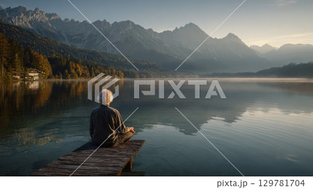 A peaceful senior woman meditates on a wooden pier by a serene lake, surrounded by majestic mountains and a tranquil, natural landscape at sunrise or sunset 129781704