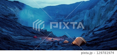 Tents are illuminated under a starry blue sky at night, camping beside a glowing, mystical blue lake within a volcanic crater 129781974