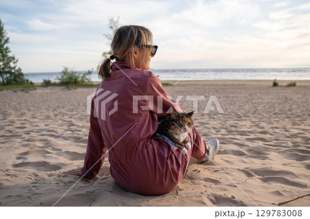 Woman relaxing on sandy beach with cat on leash at morning walk rear view. Feline curious about sand Woman relaxing on sandy beach with cat on leash at morning walk rear view. Feline curious about sand 129783008