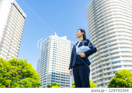 新緑の街の前にヘルメットを持って立つ女性ビジネスパーソン 129783264