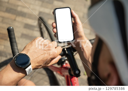 Male cyclist in helmet with mobile phone with white blank screen on bike check apps at park. Space for design. View from above. Close up. Male cyclist in helmet with mobile phone with white blank screen on bike check apps at park. Space for design. View from above. Close up. 129785386