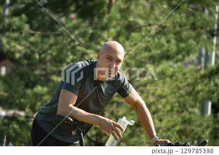 Caucasian adult cyclist tired, and relax near bicycle at greenery public park. Relaxation and refreshment outdoors among greenery. Take a break, looking at camera. Summer activity. 129785398