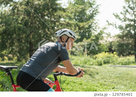 Portrait of cyclist wearing gray t-shirt in white helmet at city public park. Summer outdoor activities. Side back. Portrait of cyclist wearing gray t-shirt in white helmet at city public park. Summer outdoor activities. Side back. 129785401