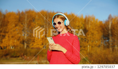 Happy young woman with phone enjoying listening to music in headphones outdoors in autumn park Happy young woman with phone enjoying listening to music in headphones outdoors in autumn park 129785837
