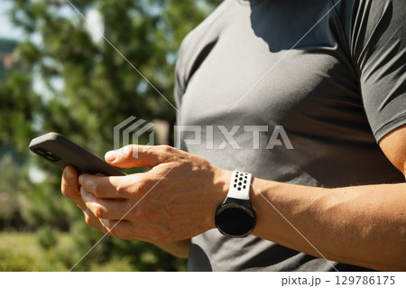 Close up of athlete hands wearing gray t-shirt with smartwatch holding mobile phone. Check apps, focused on device. Fitness progress on app. 129786175