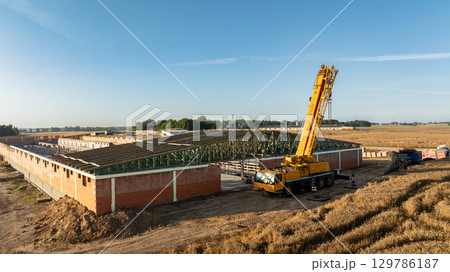 A mobile telescopic crane lifting green steel roof trusses onto a large brick structure in a rural construction site, surrounded by wheat fields and clear blue sky in early morning light 129786187