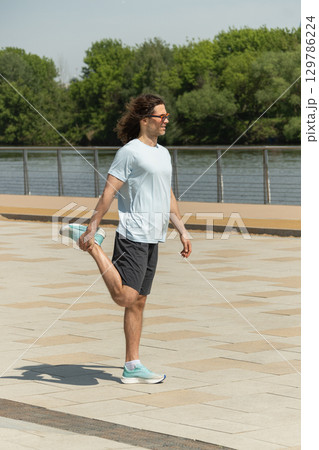 Fit handsome brunette man wearing sports casual clothes and sunglasses doing warm up exercise on city modern outdoor public park. Pause in work. Summer activity. Healthy lifestyle. 129786224